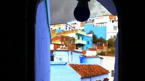 A view through a window with a blue exterior, revealing a densely built urban area with closely packed buildings. - Riad Antek (Chefchaouen)