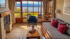A cozy living room with a fireplace, a sofa, a dining table with a bottle and glasses, and a view of mountains and a lake. - Pailahue Lodge & Cabañas (San Carlos de Bariloche)
