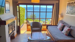 A cozy room with a sofa, a chair, a wooden table, and a large window offering a view of mountains and a lake. - Pailahue Lodge & Cabañas (San Carlos de Bariloche)