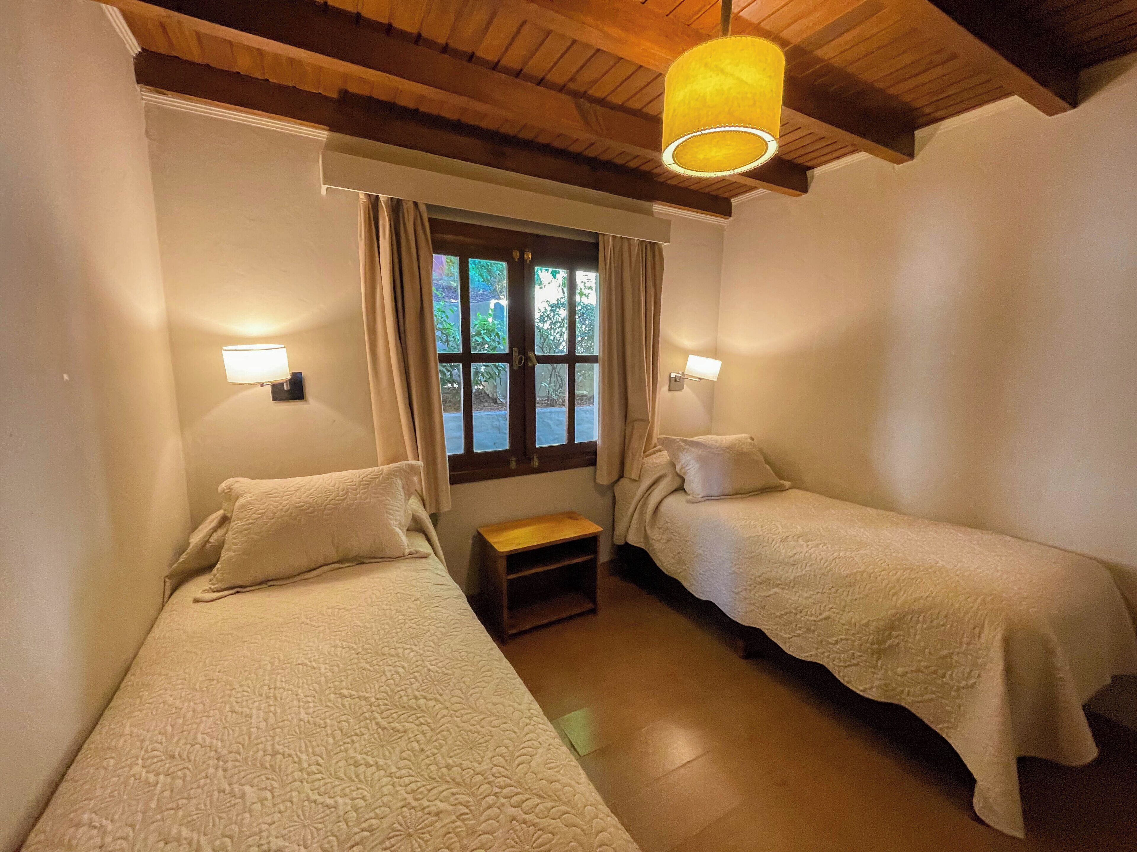 Een kamer met twee bedden, een raam met gordijnen, een houten plafond en wandlampen.