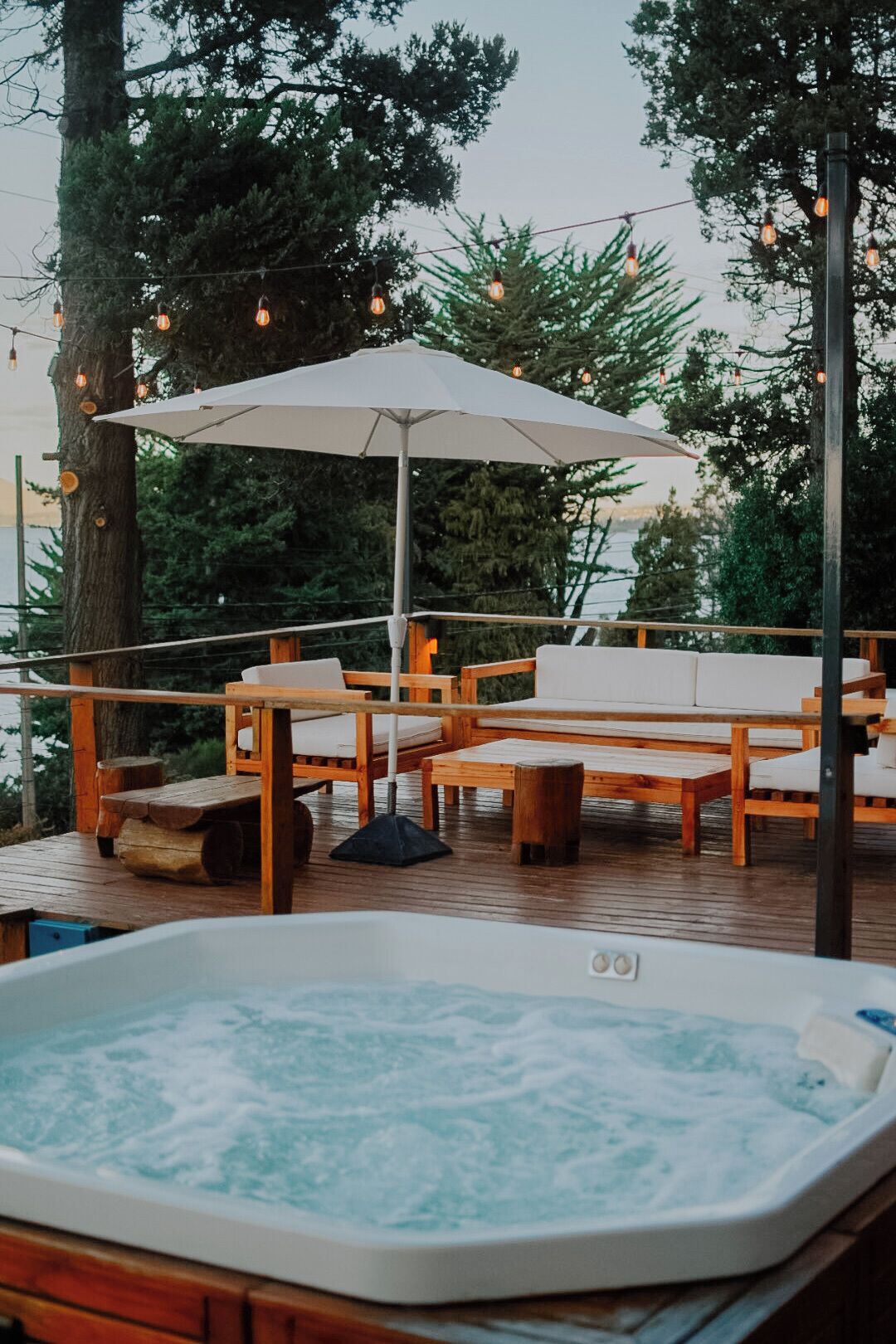 A hot tub surrounded by wooden deck chairs and an umbrella, set in a wooded area with trees and string lights.