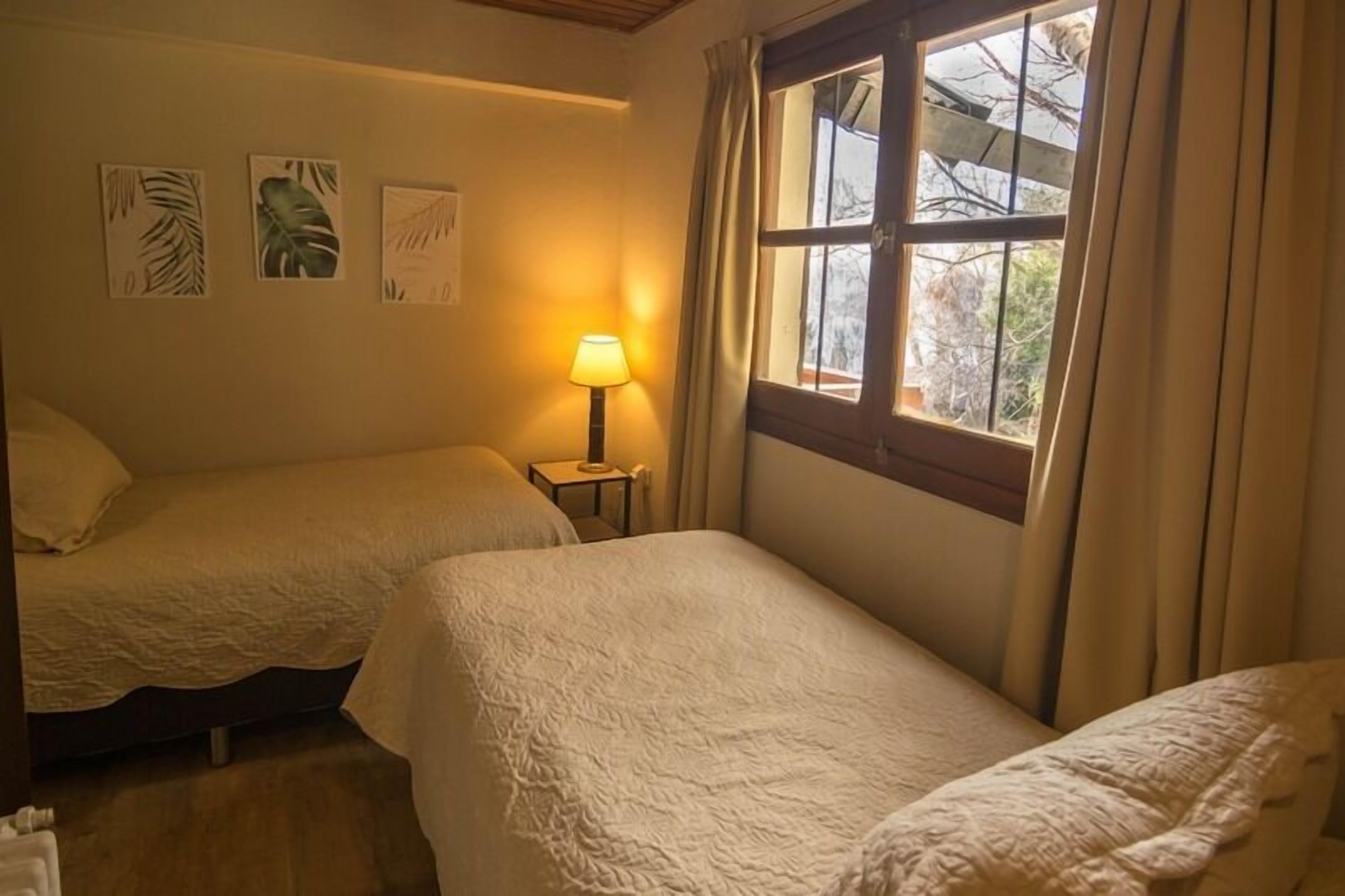 A room with two beds, a bedside table with a lamp, and a window with curtains.
