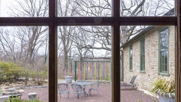 Blick durch ein Fenster auf einen Patio mit einem Steinhaus, Gartenmöbeln und einer Pergola.
