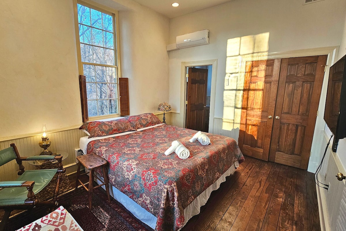 A bedroom with a large bed, wooden floor, a window with shutters, an air conditioning unit, and a door.