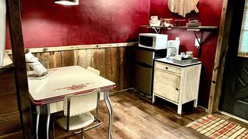 Een kleine keuken met een houten tafel en stoelen, een magnetron, een koelkast, en een klein plankje met keukengerei.