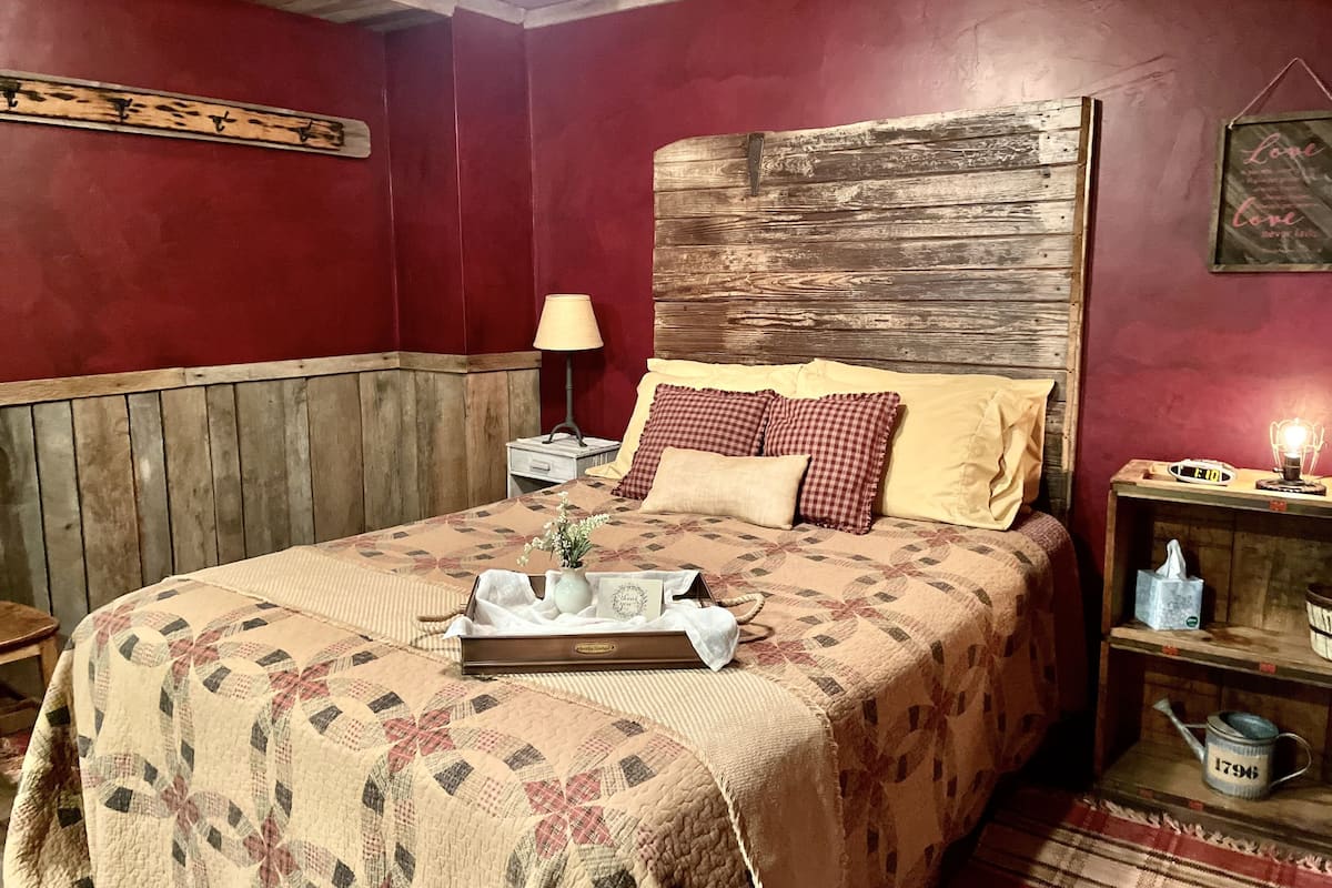 Ein gemütliches Schlafzimmer mit einem Holzkopfende, einer gesteppten Bettdecke, einem Nachttisch mit Lampe und einem Beistelltisch mit einer Uhr.