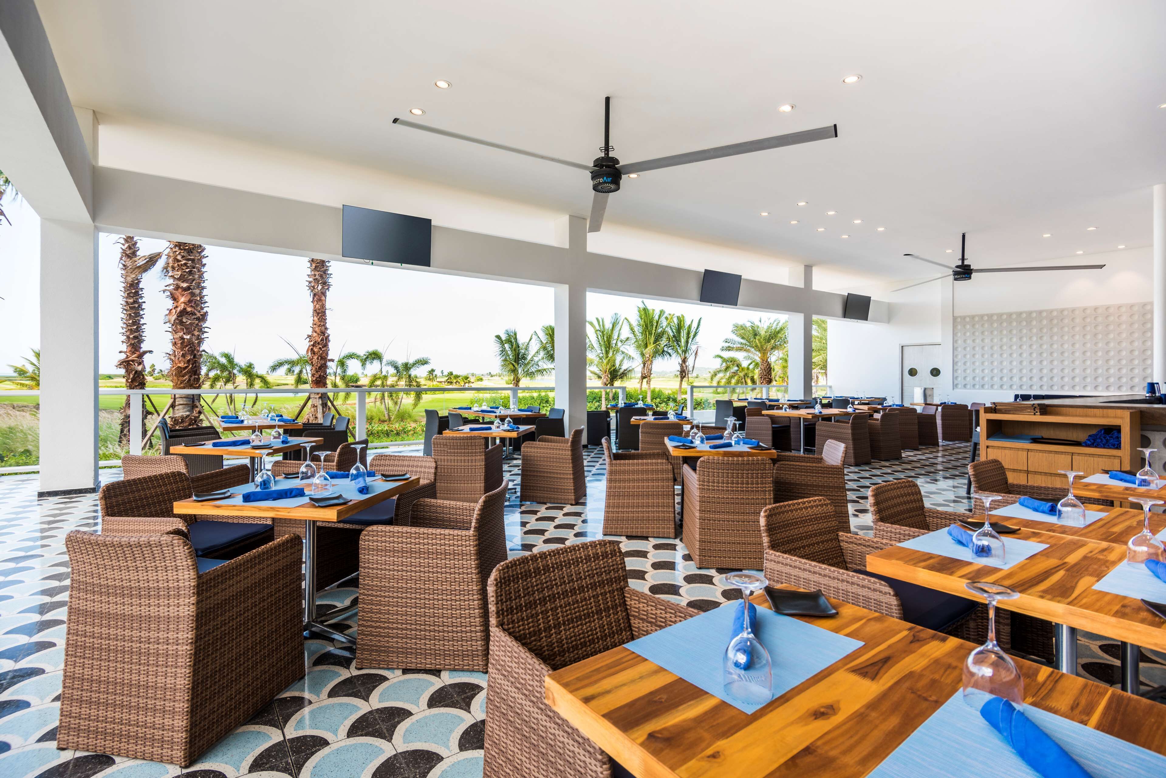 A modern outdoor dining area with wicker furniture and wooden tables, large windows offering views of palm trees and a golf course.
