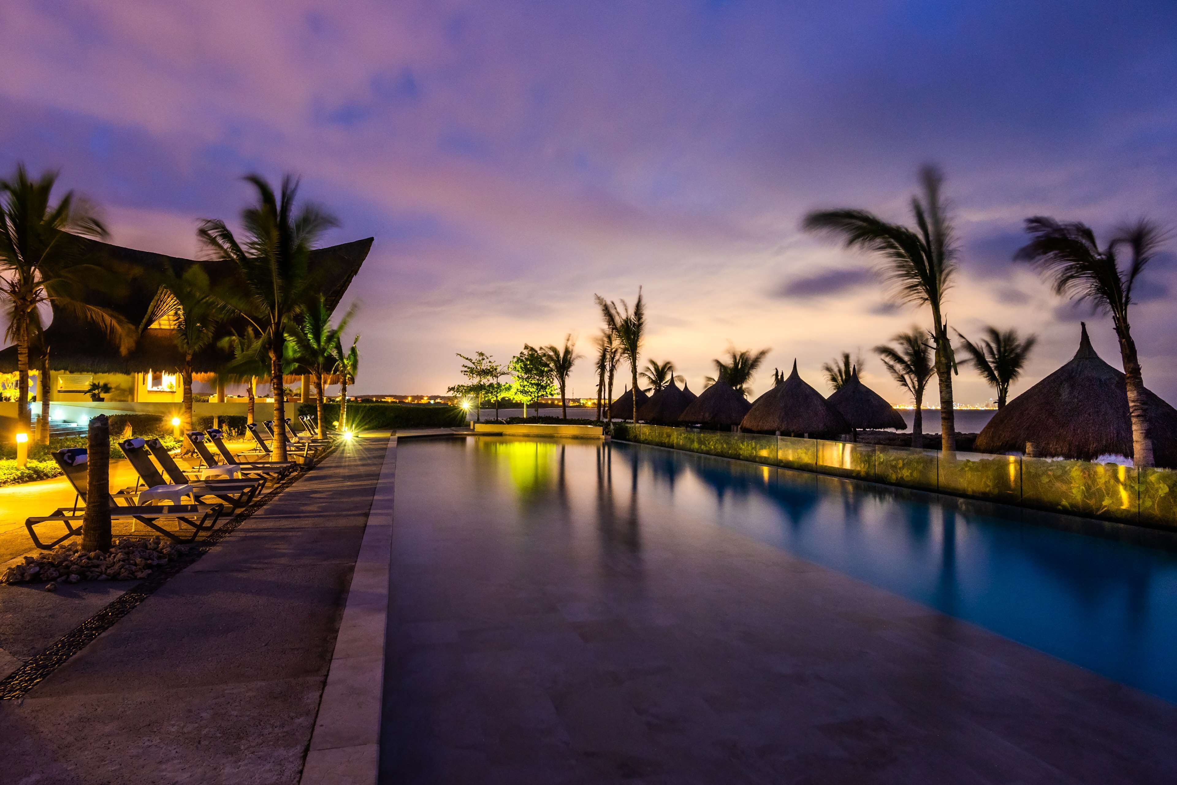 A poolside area with lounge chairs, palm trees, and thatched-roof huts.