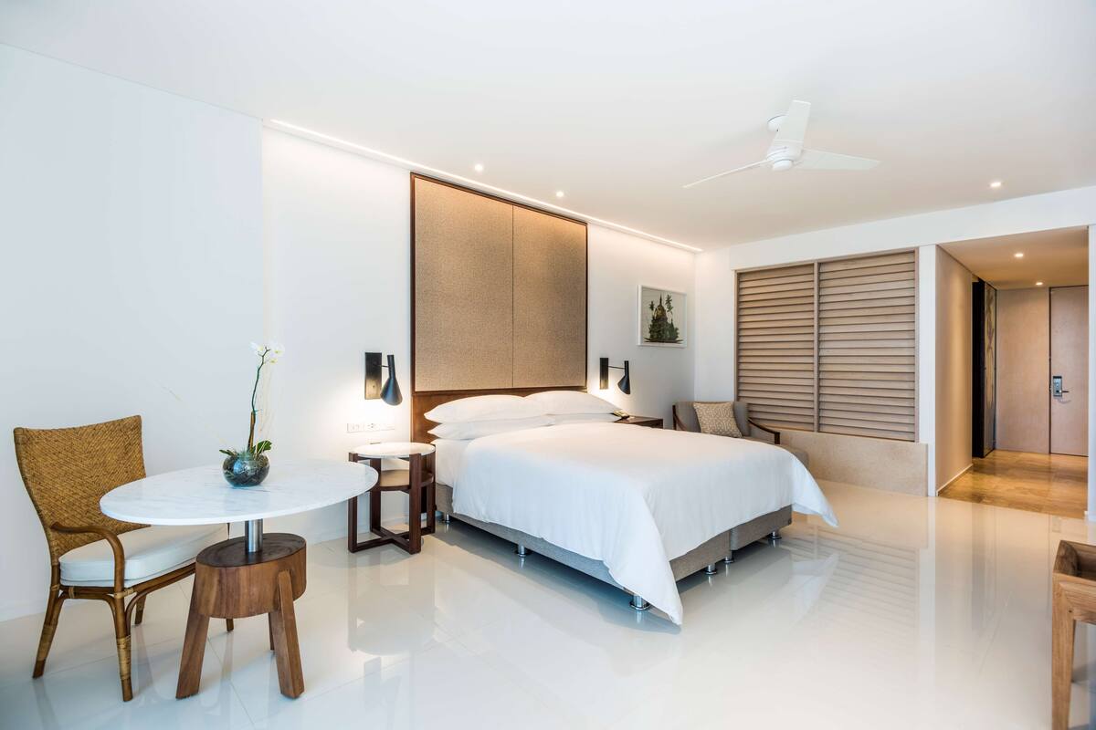 Une chambre d’hôtel moderne avec un grand lit, une petite table ronde, une chaise et un ventilateur de plafond.