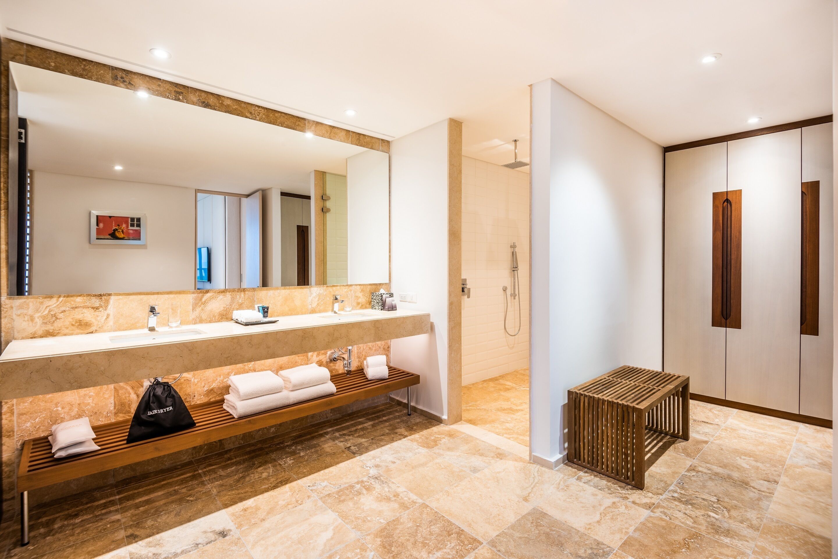 A modern bathroom with a large mirror, dual sinks, a shower, and a wooden bench.