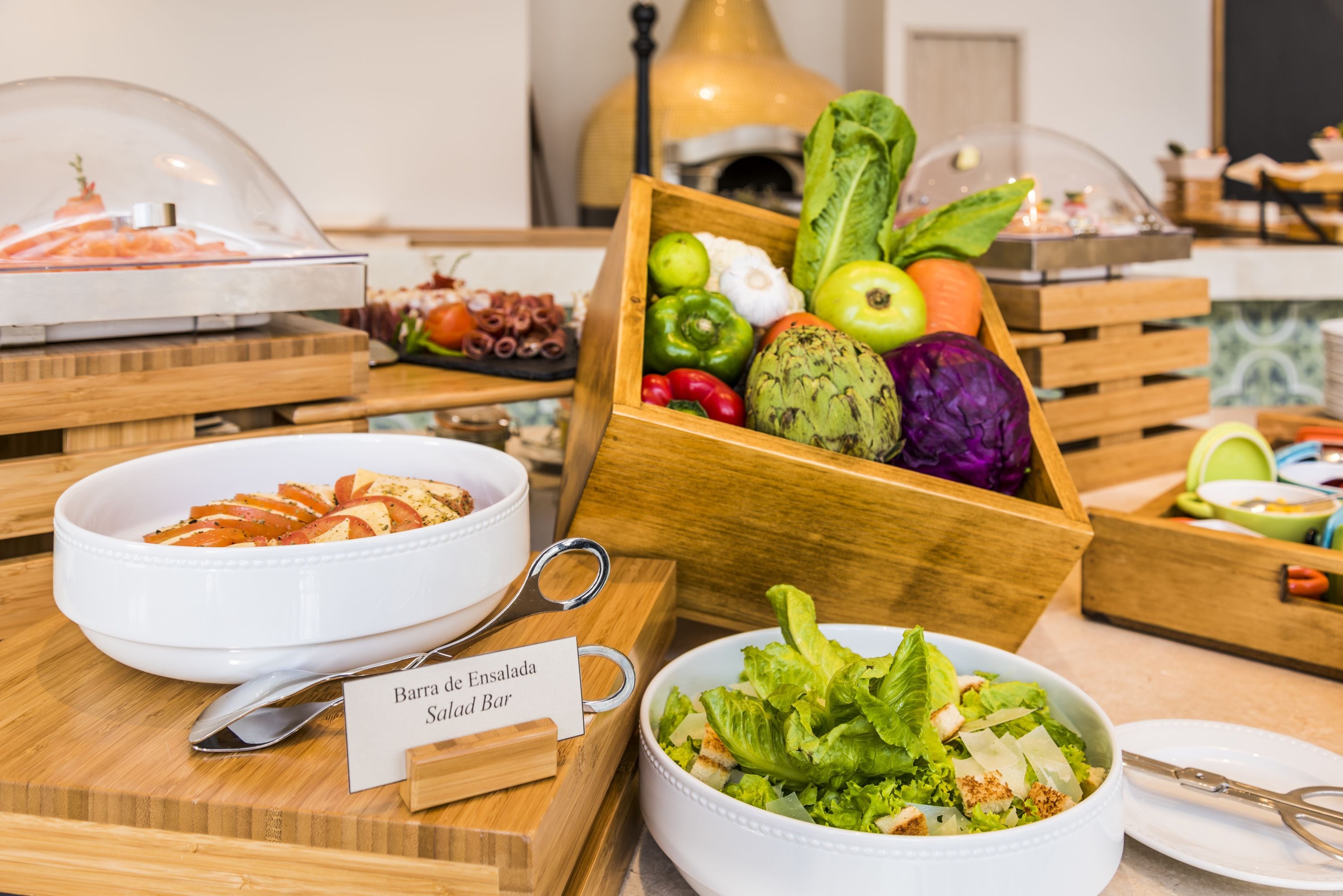 A buffet with various dishes including a bowl of sliced tomatoes, a bowl of salad, and a wooden box containing fruits and vegetables.