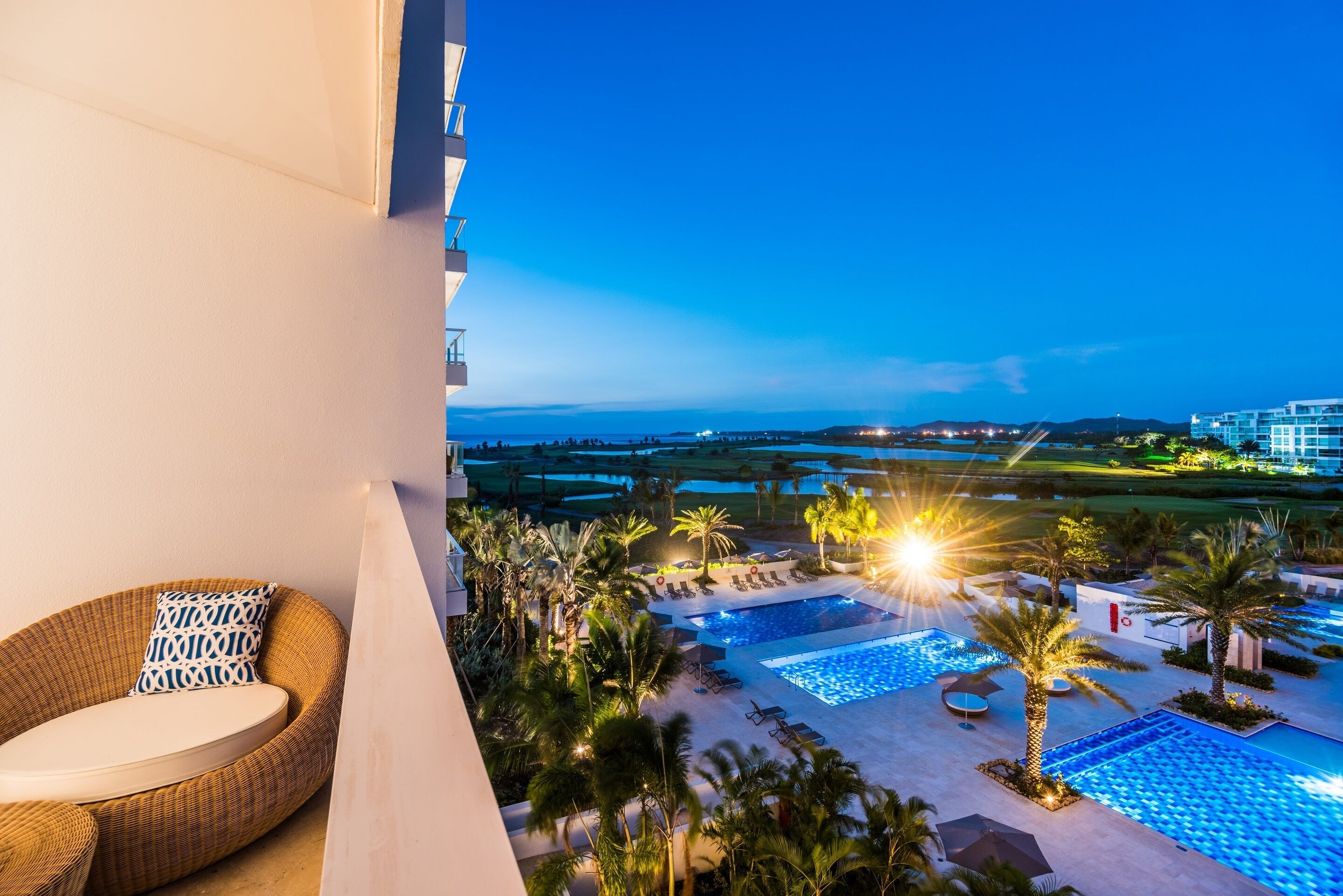 A balcony with a wicker chair and a view of a resort pool area at dusk.