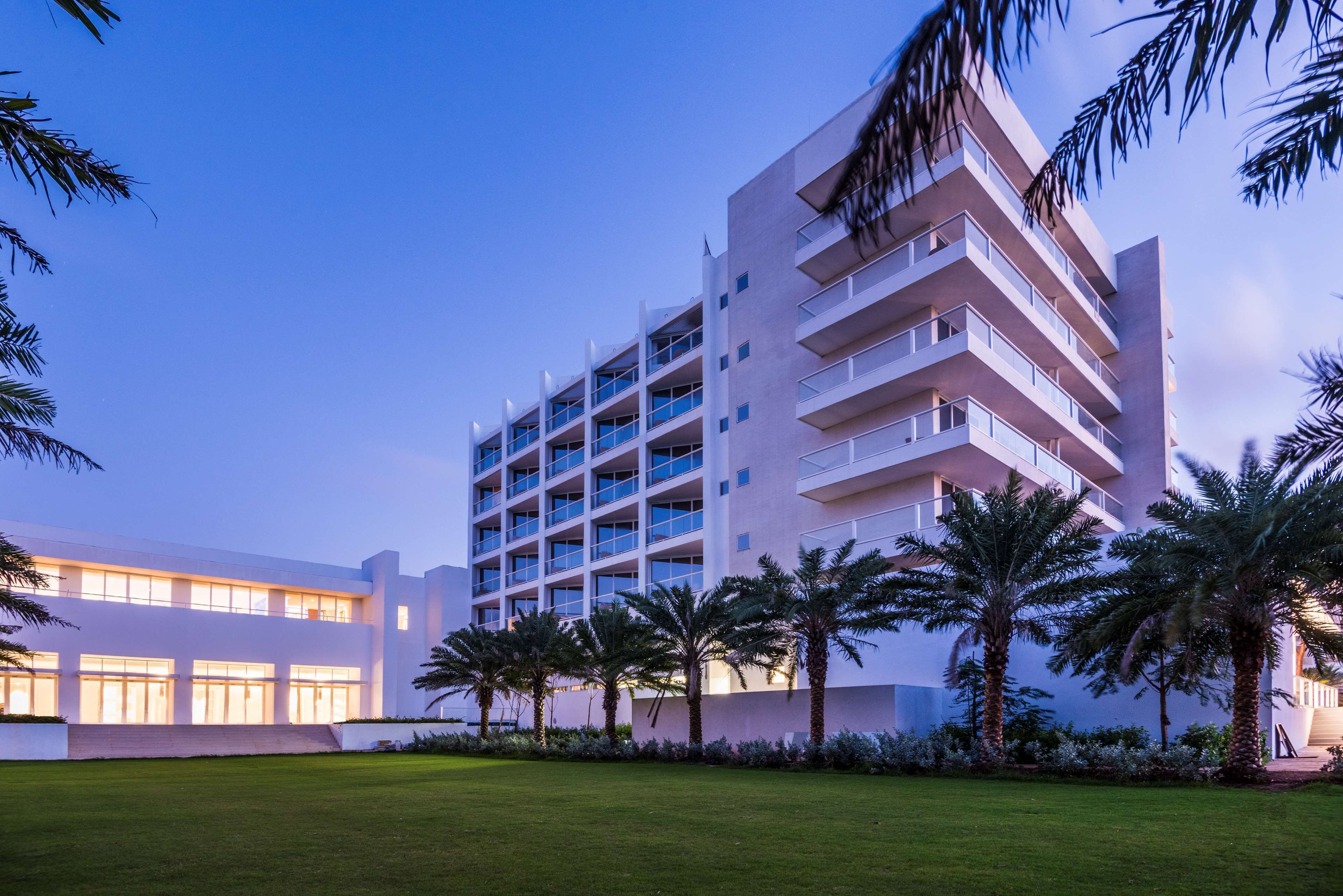 A multi-story hotel with a modern architectural design, featuring balconies and a well-maintained lawn with palm trees.