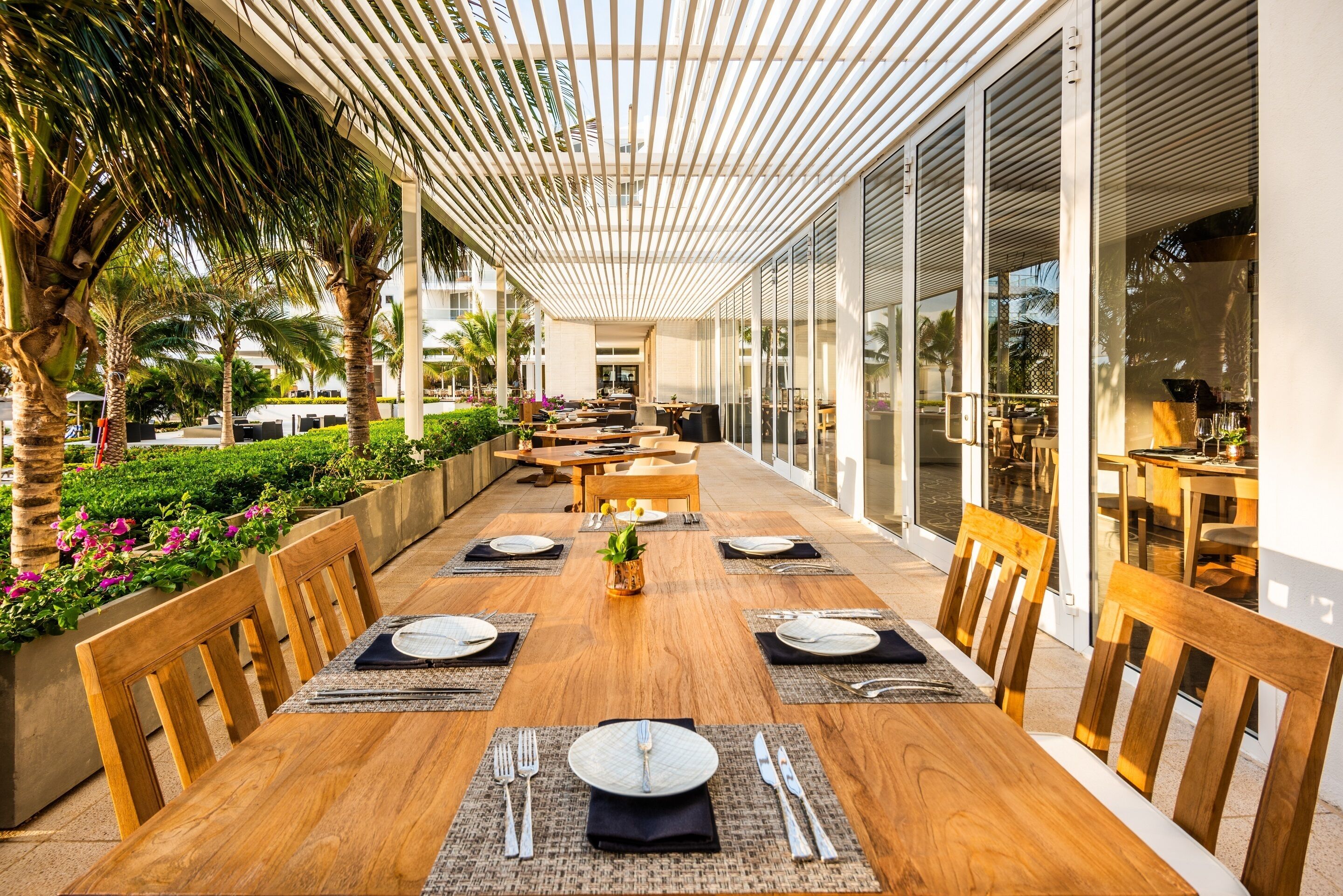 A dining area with a long wooden table set for six, surrounded by wooden chairs, with a glass partition on one side and a view of the outdoors.