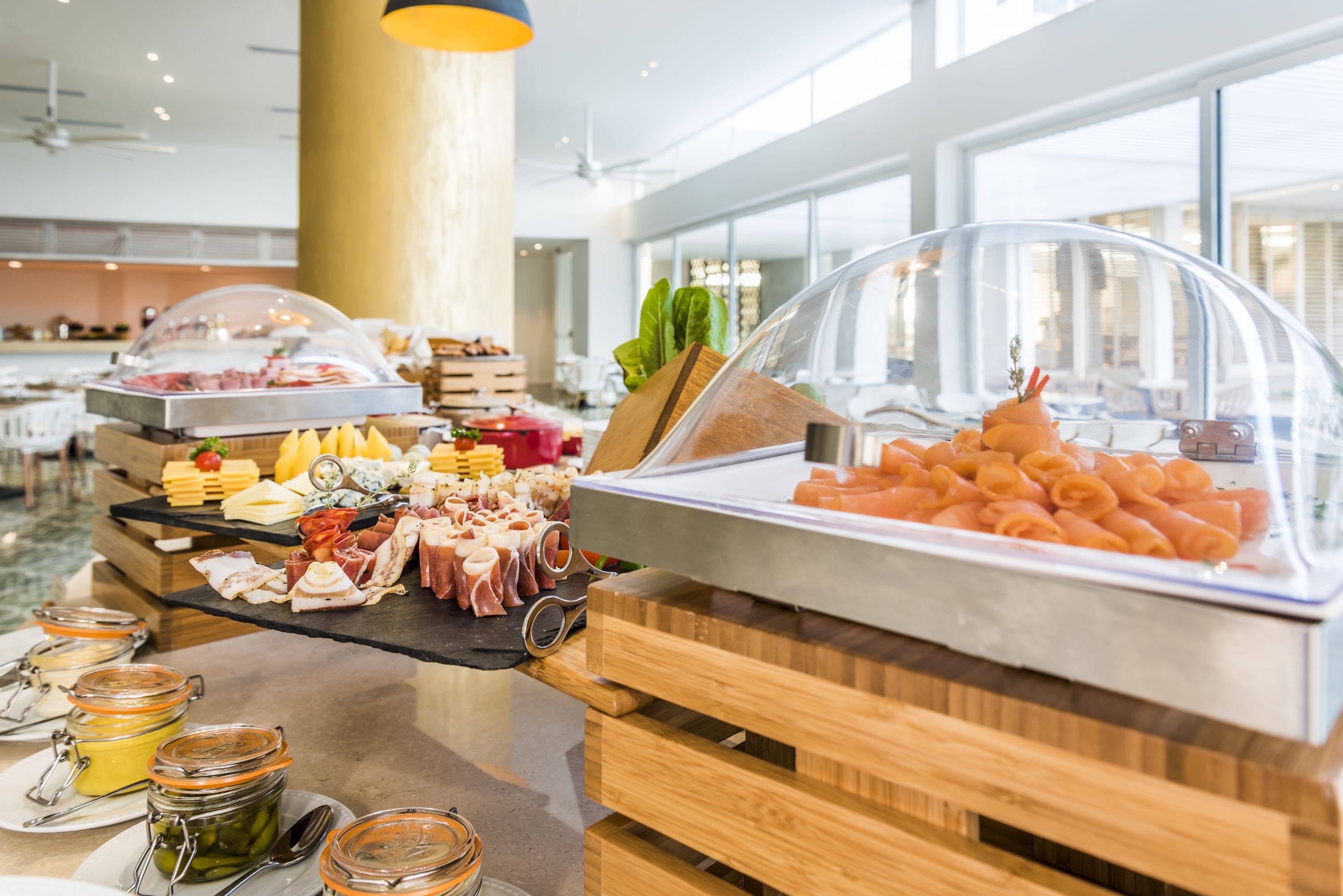 En buffet med forskellige retter, herunder kød, oste og grøntsager, præsenteret på træborde og under glaslåg.