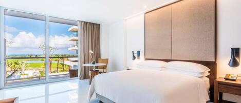 A modern hotel room with a large bed, a balcony with a view of the ocean, a round table, and a ceiling fan.