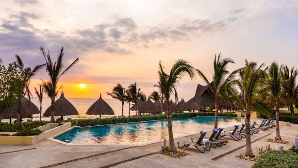Een zwembadgedeelte van een resort met uitzicht op de zonsondergang, rieten parasols en ligbedden.