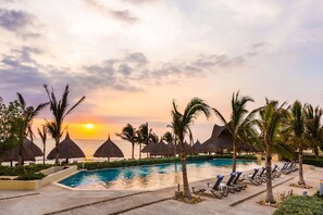Área de piscina en un resort con vista al atardecer, sombrillas de techo de paja y sillones de descanso.