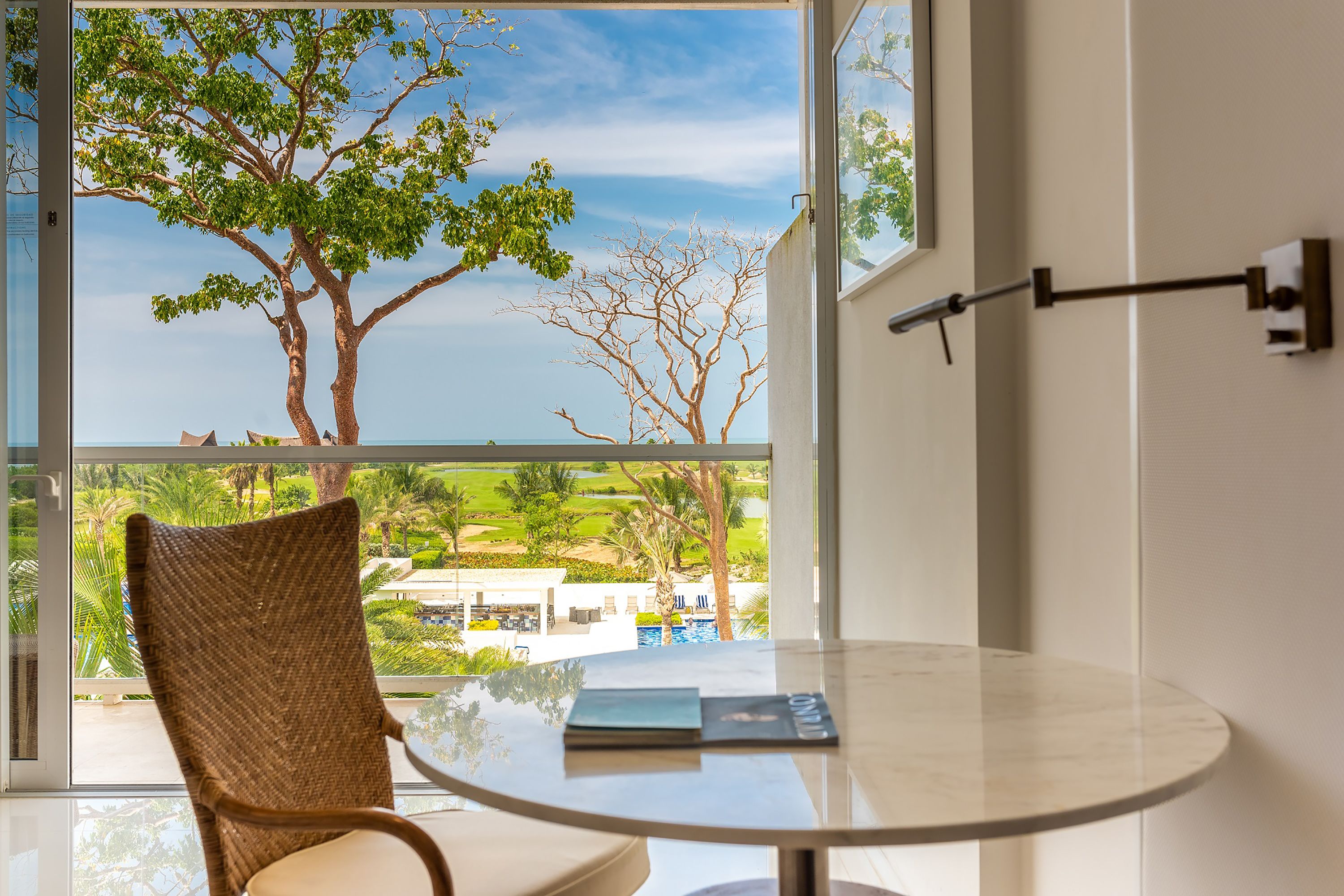 Une pièce avec une table en verre, une chaise en osier et une vue sur un arbre et un paysage, le tout visible à travers une grande fenêtre.