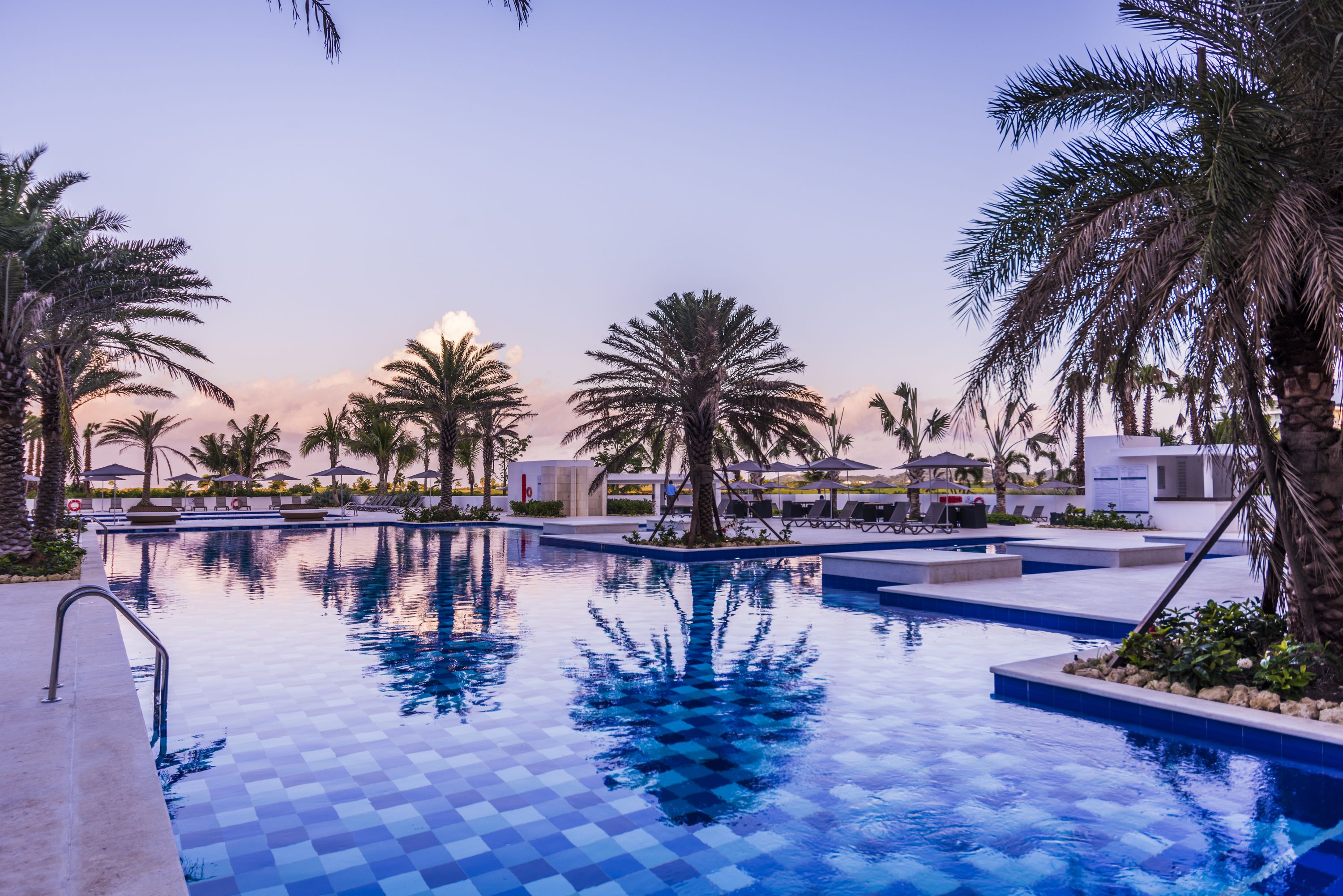 Un área de piscina con suelo de baldosas, palmeras y sillones de descanso.