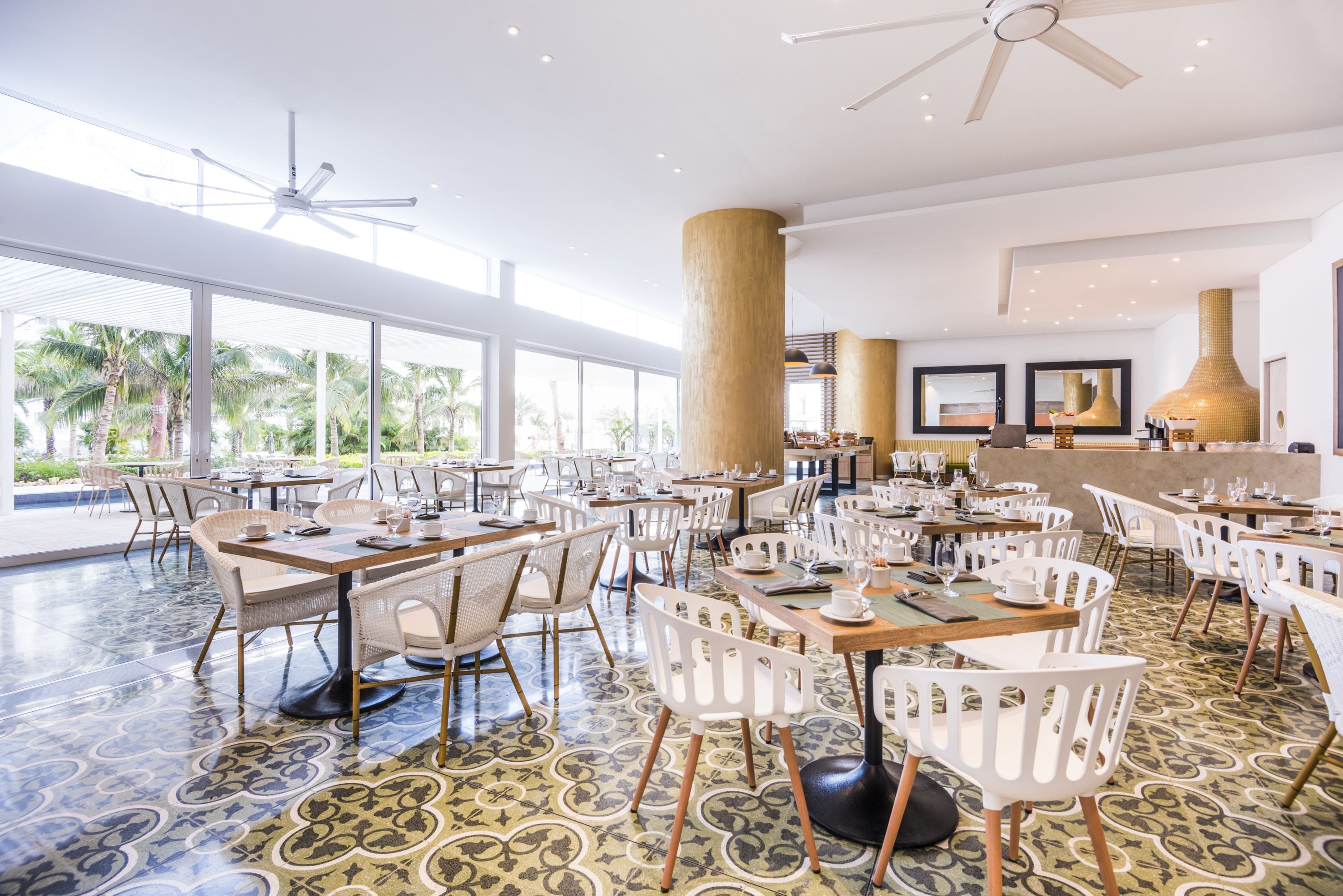 A spacious dining area with multiple tables and chairs, large windows offering a view of palm trees, and a ceiling fan.