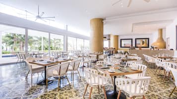 A spacious dining area with multiple tables and chairs, large windows offering a view of palm trees, and a ceiling fan.