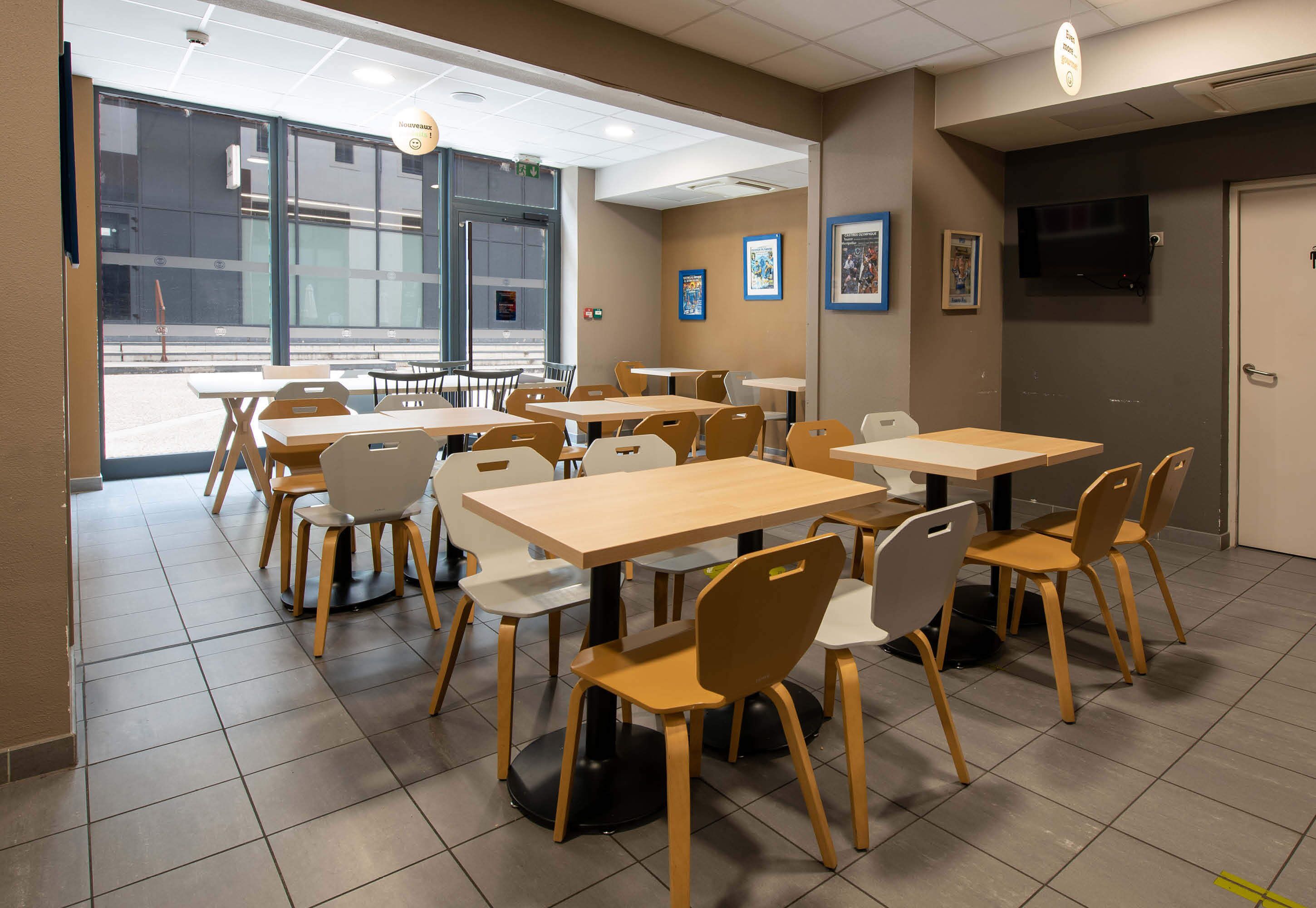 A cafeteria with multiple tables and chairs, large windows, and a television on the wall.