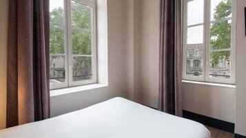 Habitación de hotel con una cama grande, dos ventanas con cortinas y vistas a edificios.