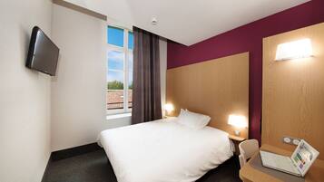 Een hotelkamer met een bed, een bureau met een laptop, een aan de muur bevestigde televisie en een raam met gordijnen.