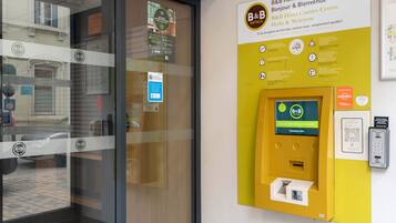 Ontvangstbalie van een B&B Hotel met een glazen deur, een geel informatiebord en een betaalautomaat.