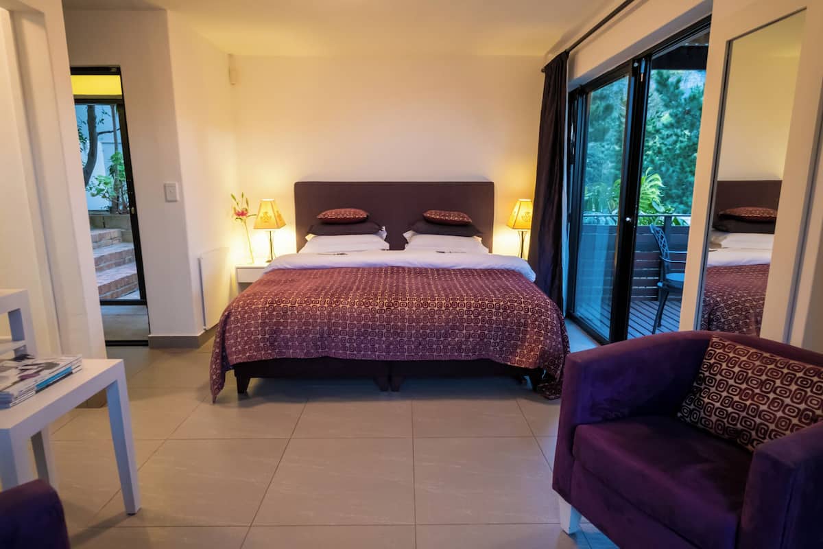 Habitación de hotel con una cama grande, dos lámparas de noche, un sofá morado y vistas al exterior a través de un ventanal corredero.
