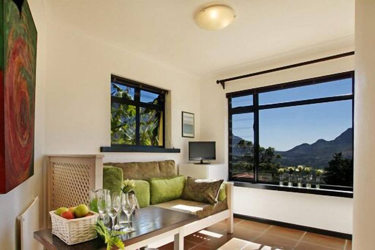 Una habitación con un ventanal amplio, un sofá, una mesa con vasos y fruta, y un cuadro en la pared.