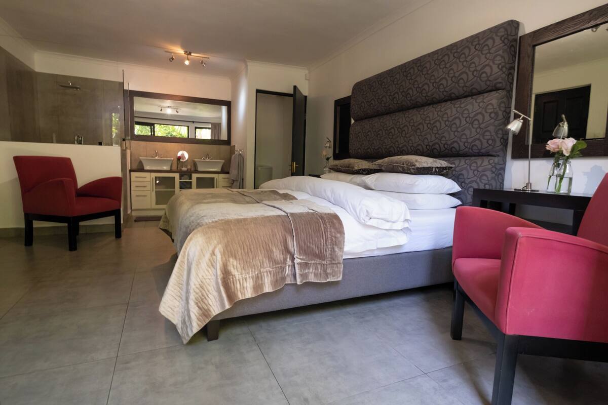 Un dormitorio moderno con una cama grande, una zona de estar con una silla, un tocador con espejo y un baño con ducha.
