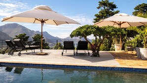 A poolside area with lounge chairs and umbrellas, a tiled pool, and a mountainous backdrop.