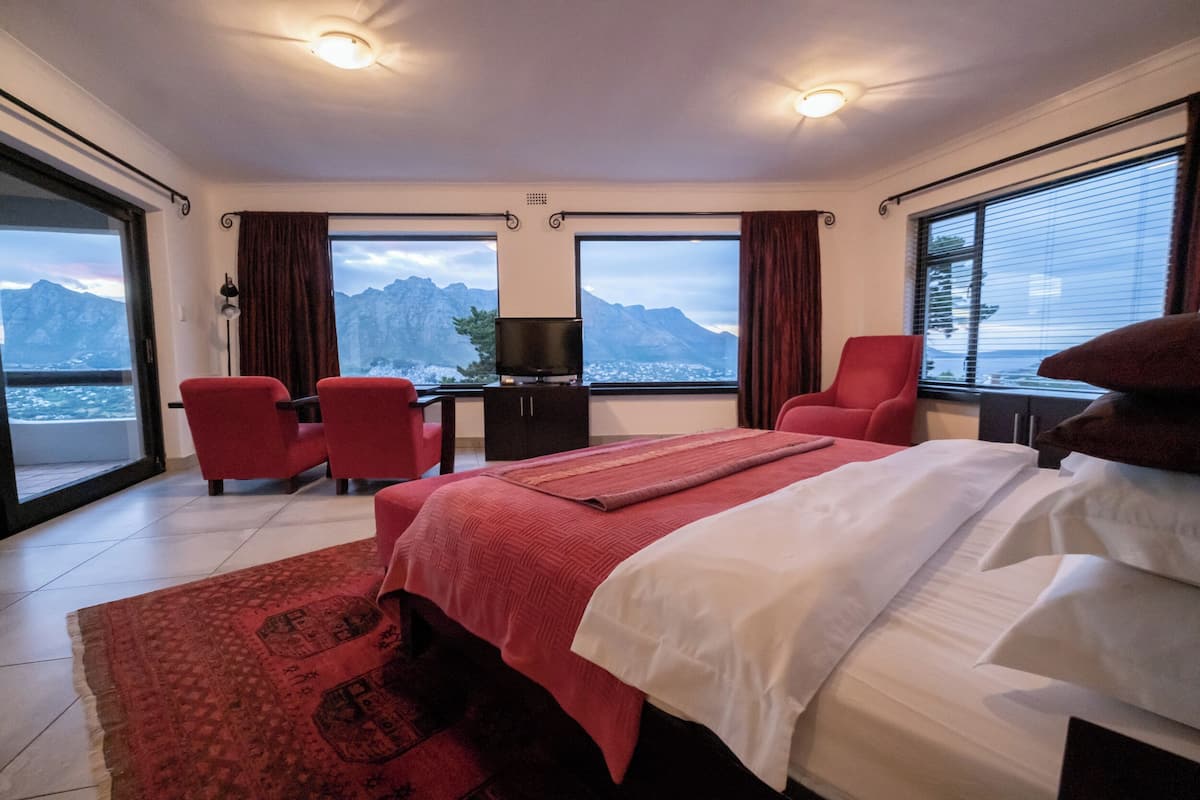 Habitación de hotel con una cama, dos sillas rojas, un escritorio con televisor y ventanas amplias con vistas a la montaña.