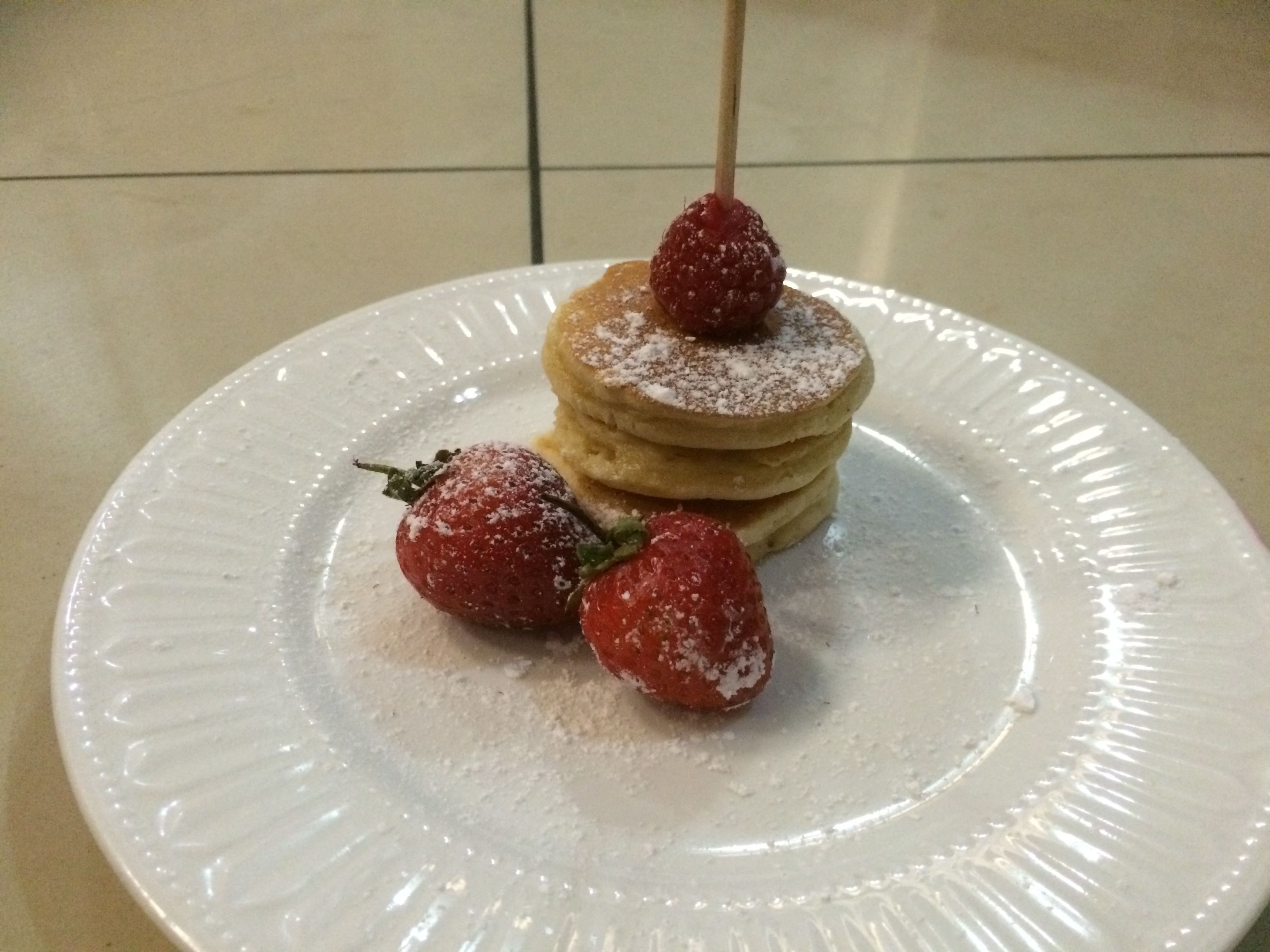Une pile de crêpes, avec une fraise sur le dessus et deux autres fraises à côté, sur une assiette blanche.