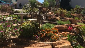 Un jardín con diversas plantas y flores, un banco de piedra y un edificio al fondo.