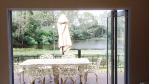 Un área de comedor al aire libre con una mesa y sillas blancas, una sombrilla y vistas a un lago y árboles a través de un ventanal.