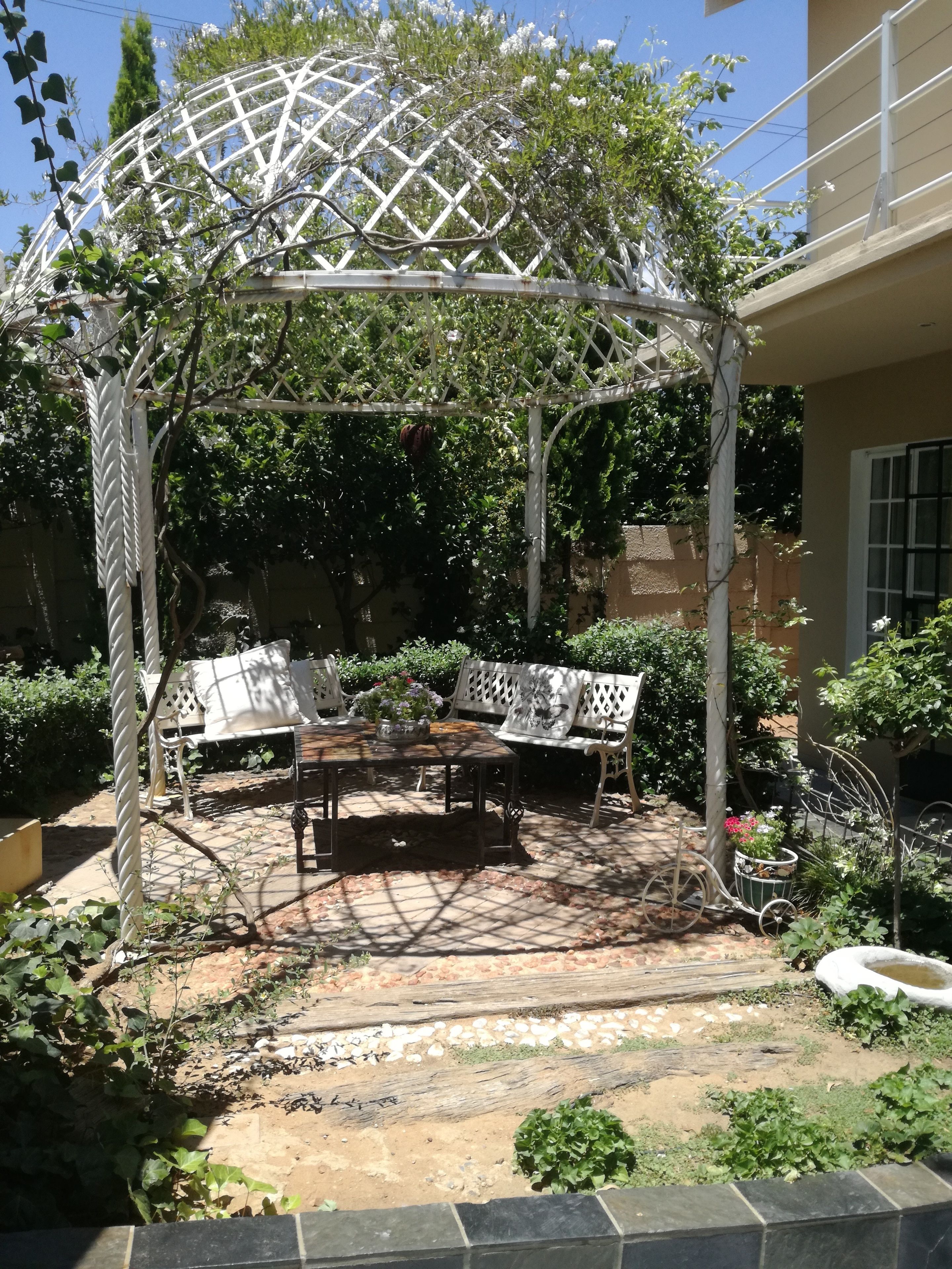 A garden patio with a pergola, two white metal chairs, a table, and various plants.