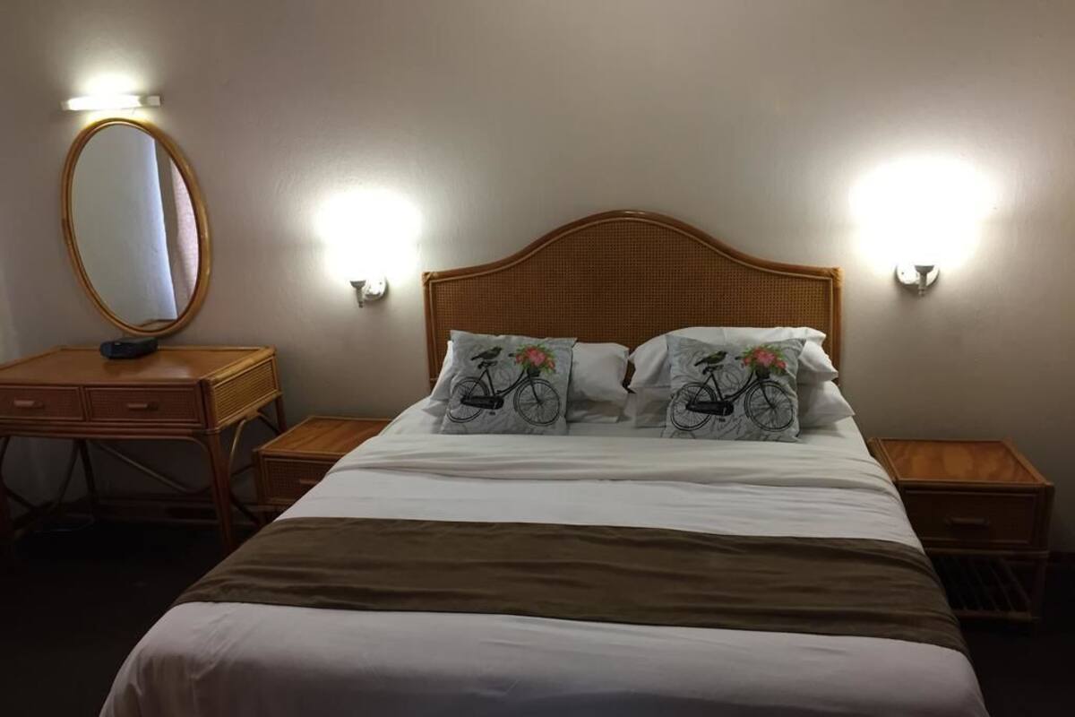 Een netjes opgemaakt bed met een houten hoofdbord, twee nachtkastjes, een spiegel en wandlampen.