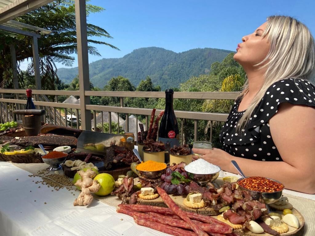 Een persoon zit aan een tafel met diverse gerechten, waaronder vlees, kaas en fruit, met een prachtig uitzicht op bergen op de achtergrond.