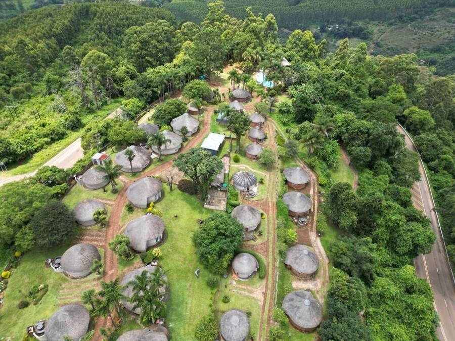 Vista aérea de um resort com várias cabanas de telhado de palha dispostas em padrão de grade, cercado por vegetação exuberante e uma estrada sinuosa.