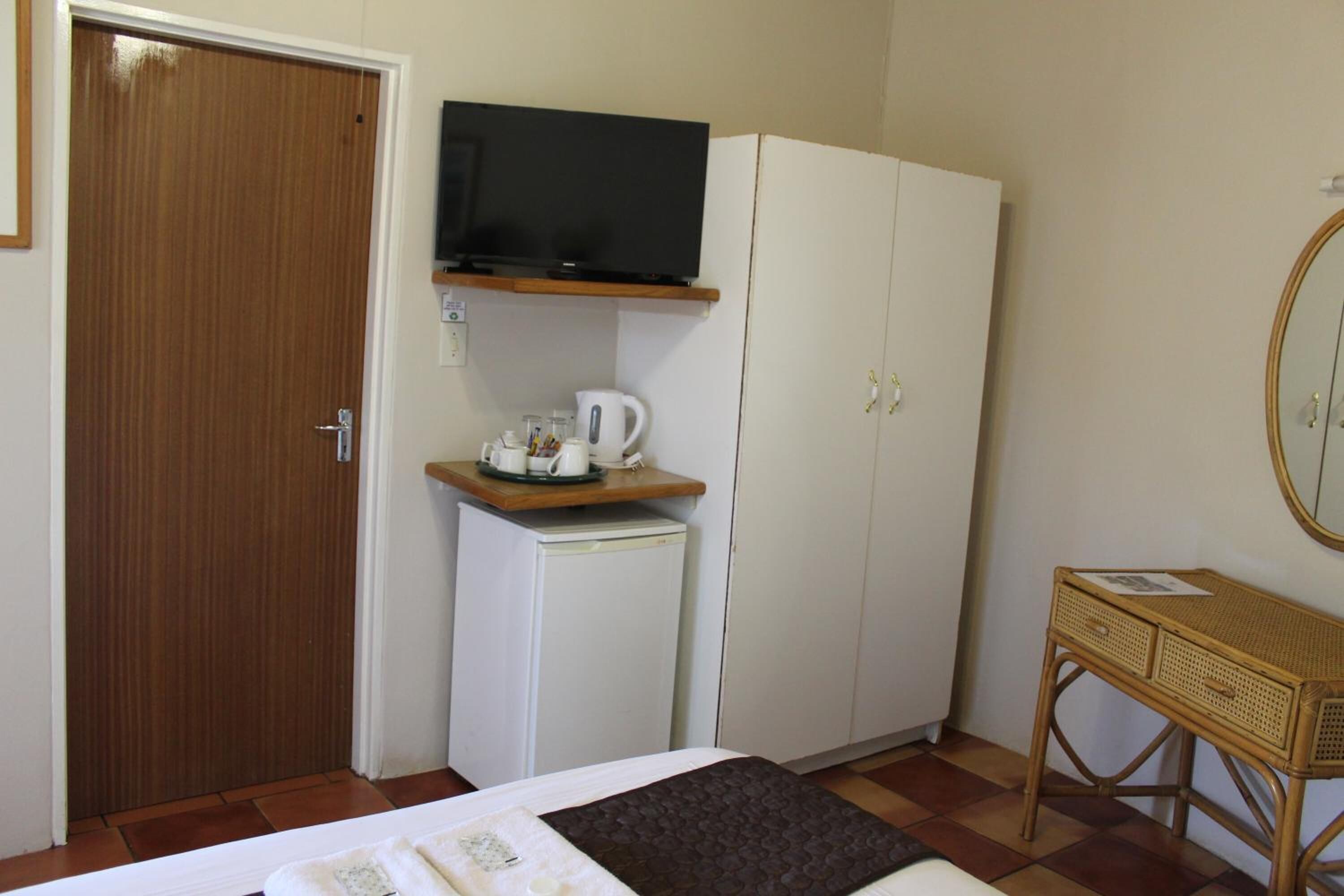 A bedroom with a bed, a television on a shelf, a small refrigerator, and a bedside table with a mirror.