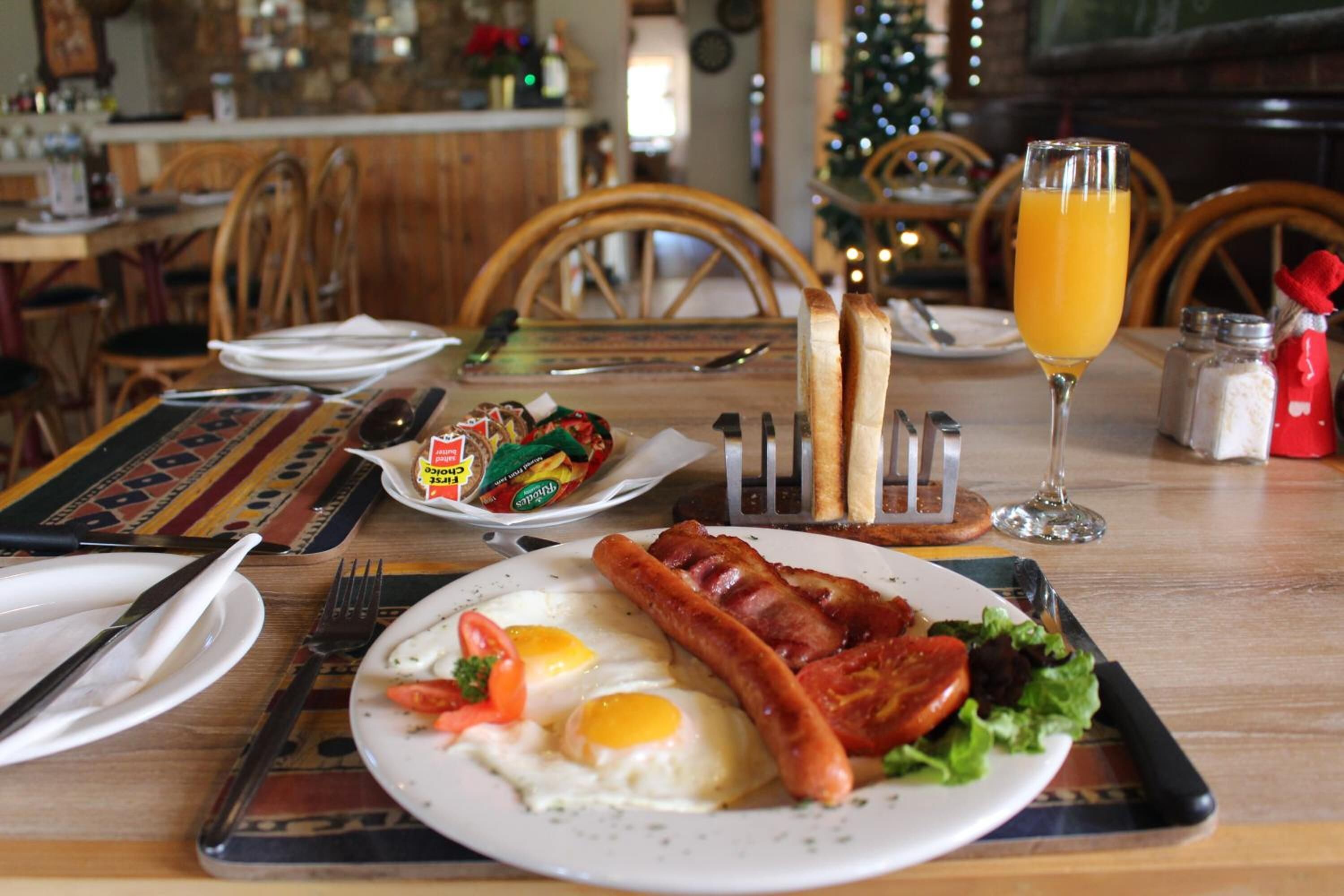 Um ambiente de café da manhã com um prato de ovos, bacon e legumes, um copo de suco de laranja, e uma árvore de Natal ao fundo.