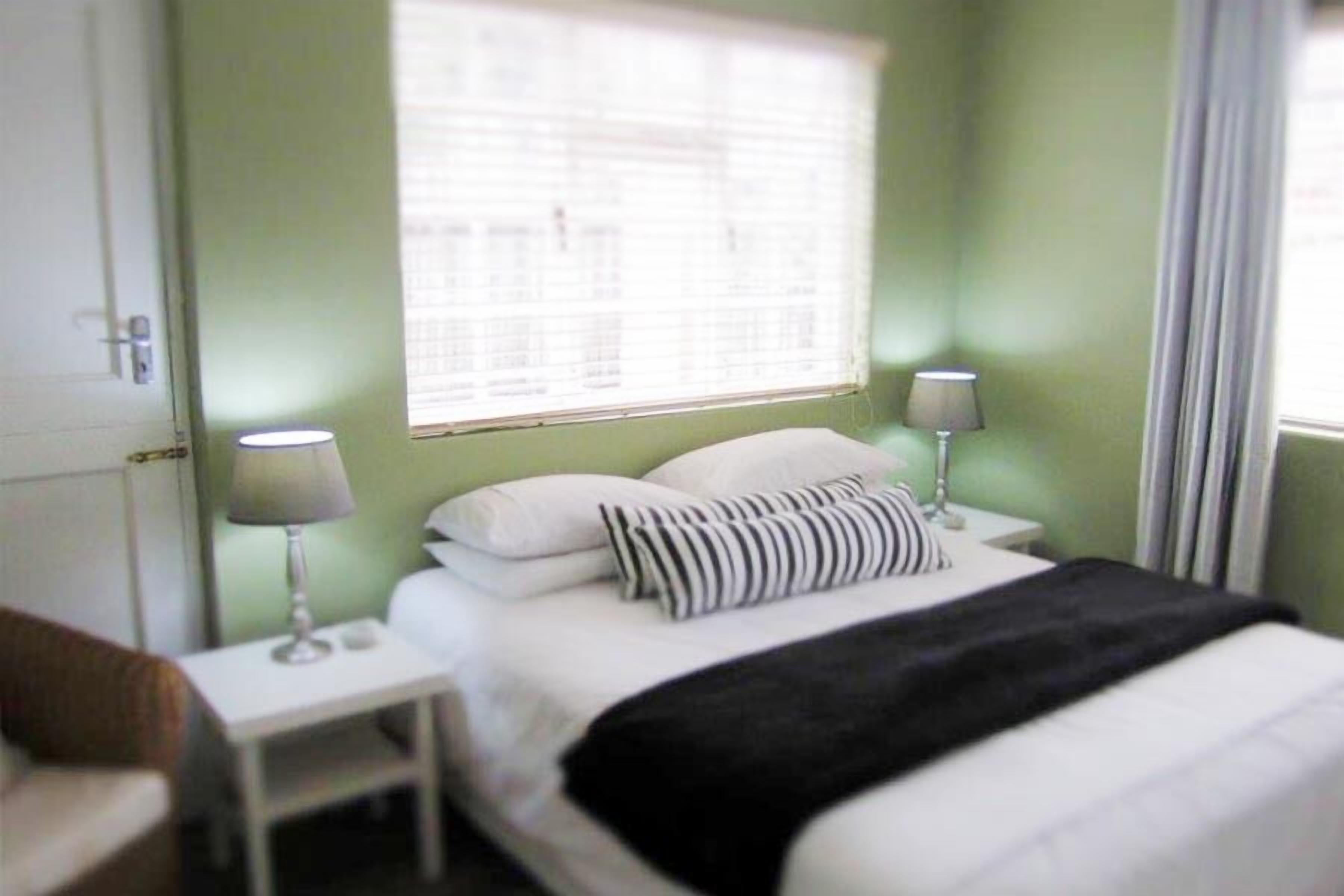 A bedroom with a bed, two bedside tables with lamps, a window with blinds, and a door.