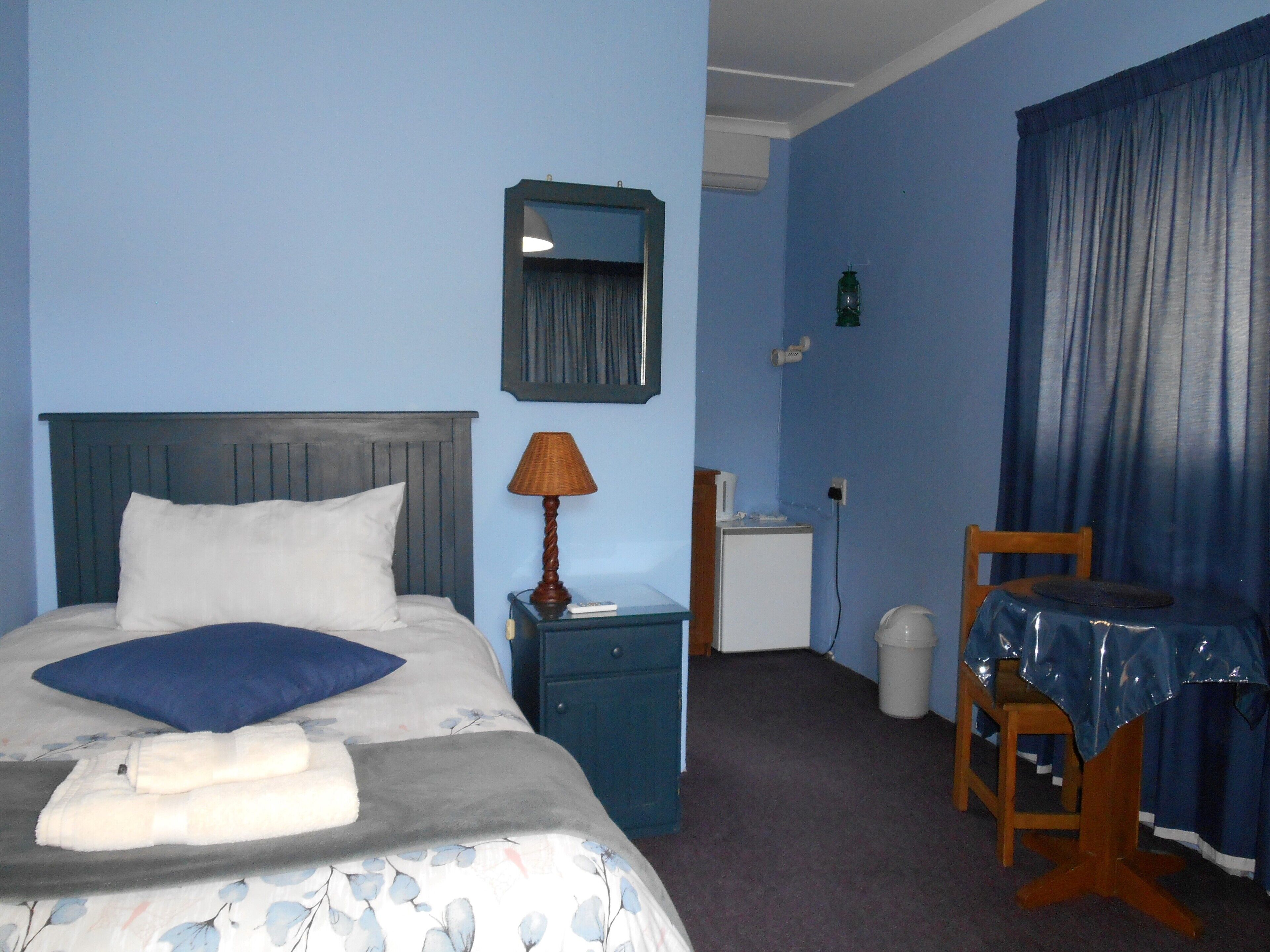 A hotel room with a bed, a nightstand, a lamp, a small table with a tablecloth, a chair, and a window with curtains.