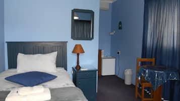 A hotel room with a bed, a nightstand, a lamp, a small table with a tablecloth, a chair, and a window with curtains.