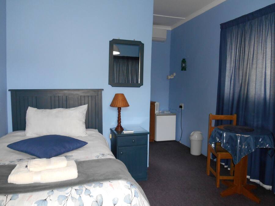 A hotel room with a bed, a nightstand, a lamp, a small table with a tablecloth, a chair, and a window with curtains.