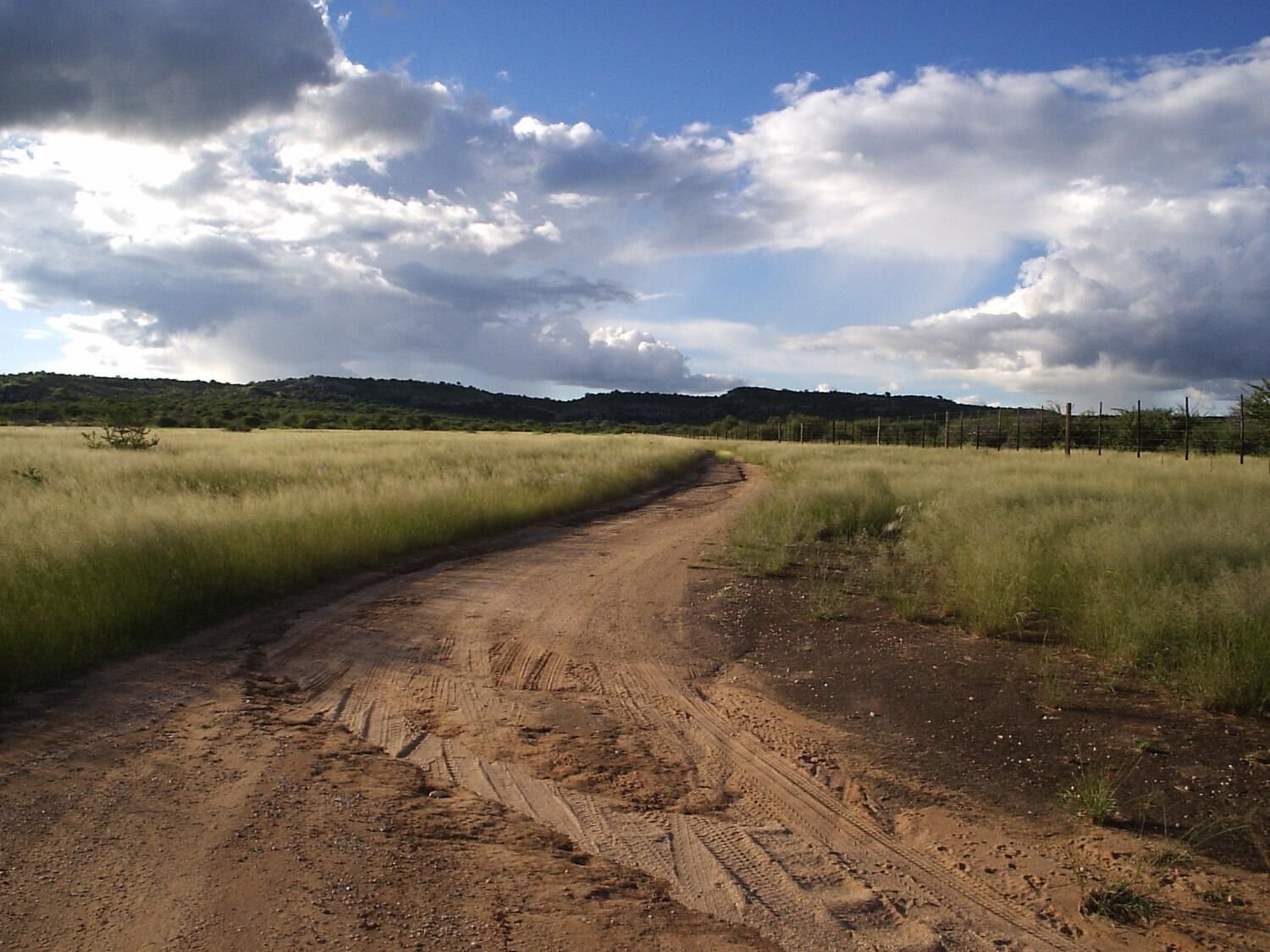 A dirt road leading through a grassy field with a fence on one side and a hill in the distance.