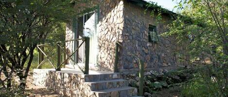 Un bâtiment en pierre à l’aspect rustique, un escalier en pierre menant à une porte, et un environnement naturel agrémenté d’arbres et d’arbustes.